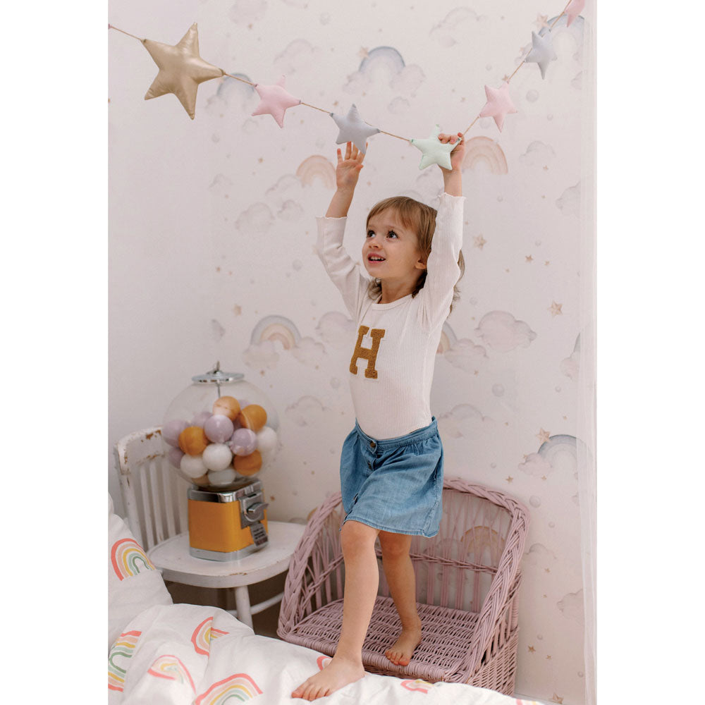 Dekornik Rainbows Behang — Een vrolijk behang met zachte regenbogen, wolkjes en sterretjes in pastelkleuren, perfect voor een vrolijke, kleurrijke kinderkamer. Sfeerbeeld van het behang in een ingerichte kinderkamer.